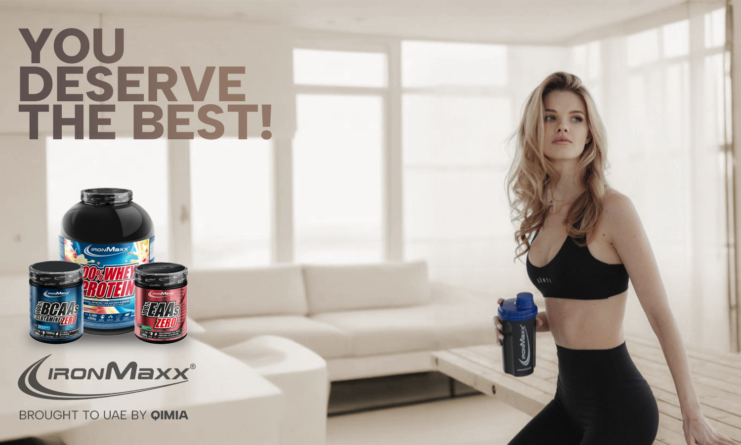 Fit woman holding IronMaxx shaker in modern room beside IronMaxx whey protein, BCAAs, and EAAs – Premium German supplements brought to UAE by Qimia Nutrition